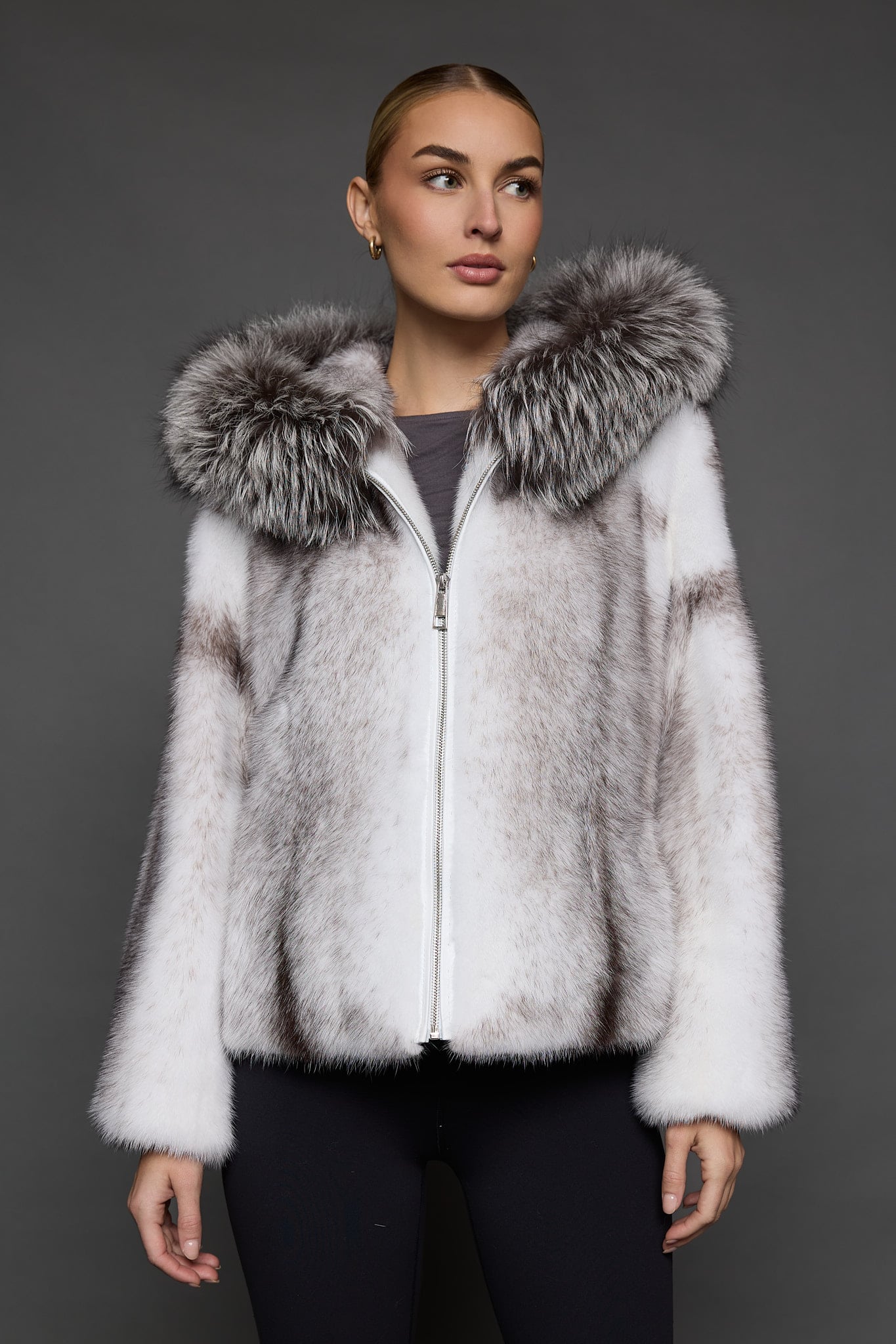 Frontal view of a woman in a black cross mink jacket with the hood down standing in front of a dark grey background