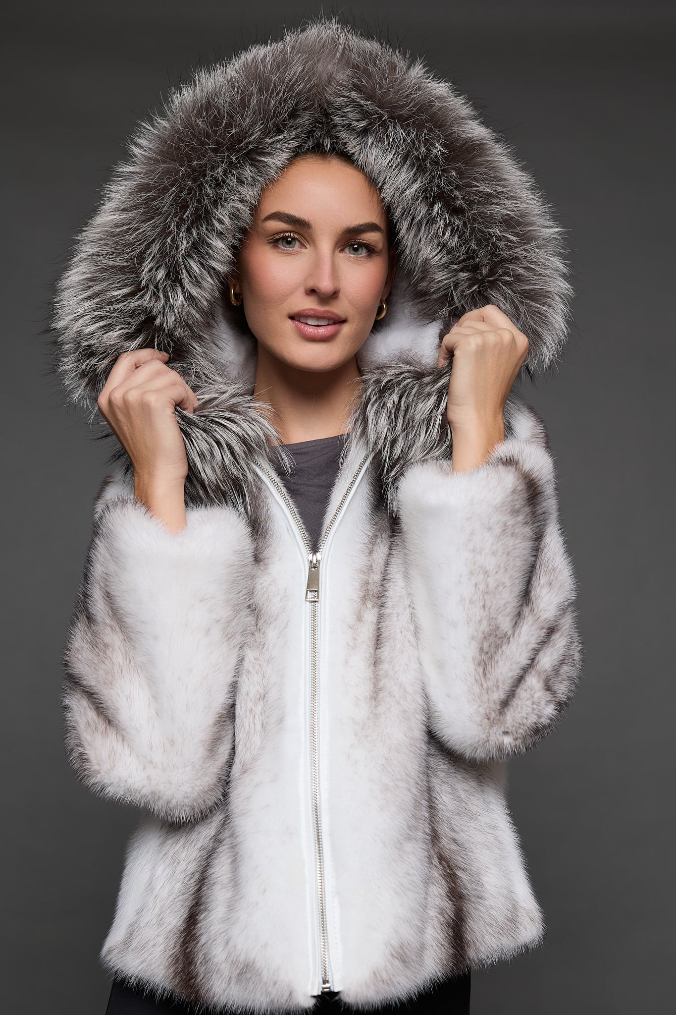 Frontal view of a woman in a black cross mink jacket with the hood up and both hands gripping the hood standing in front of a dark grey background
