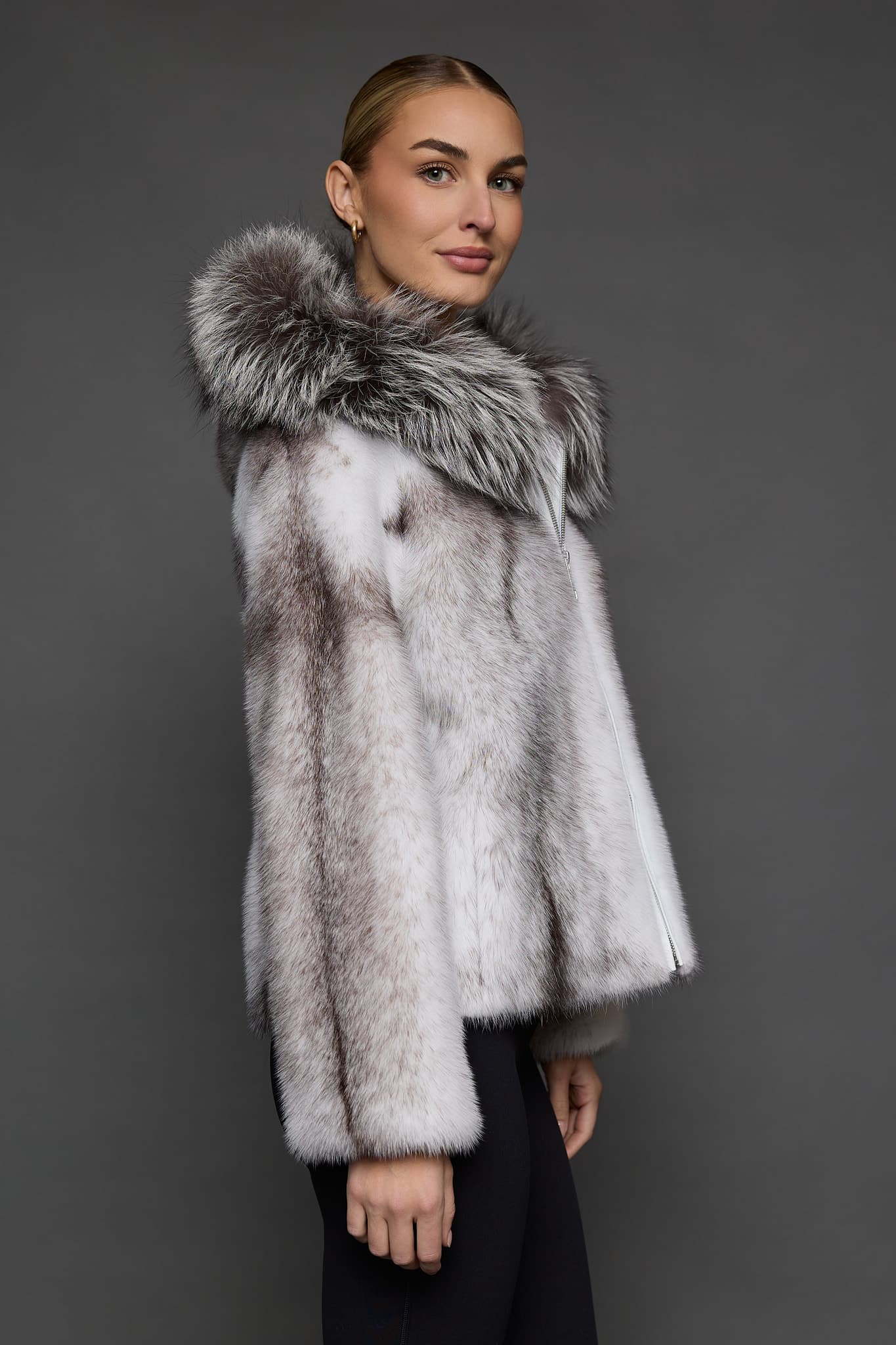 Profile view of a woman in a black cross mink jacket with the hood down looking at the camera and standing in front of a dark grey background