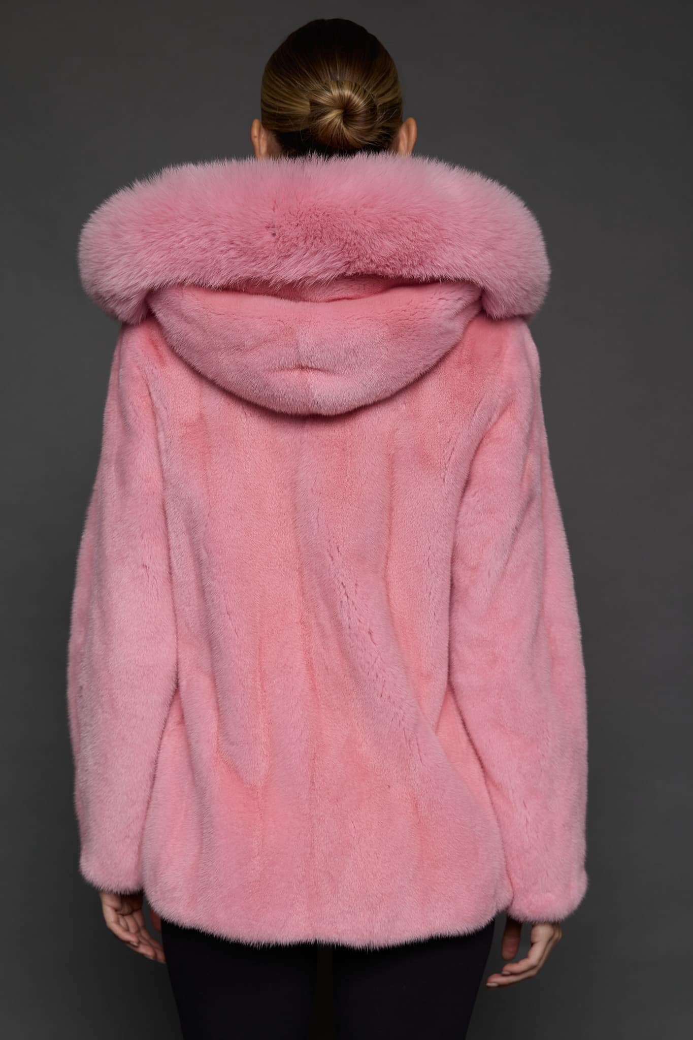 Rear view of a woman in a blush mink jacket with the hood down standing in front of a dark grey background