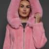 Close-up view of a woman in a blush mink jacket with the hood up standing in front of a dark grey background