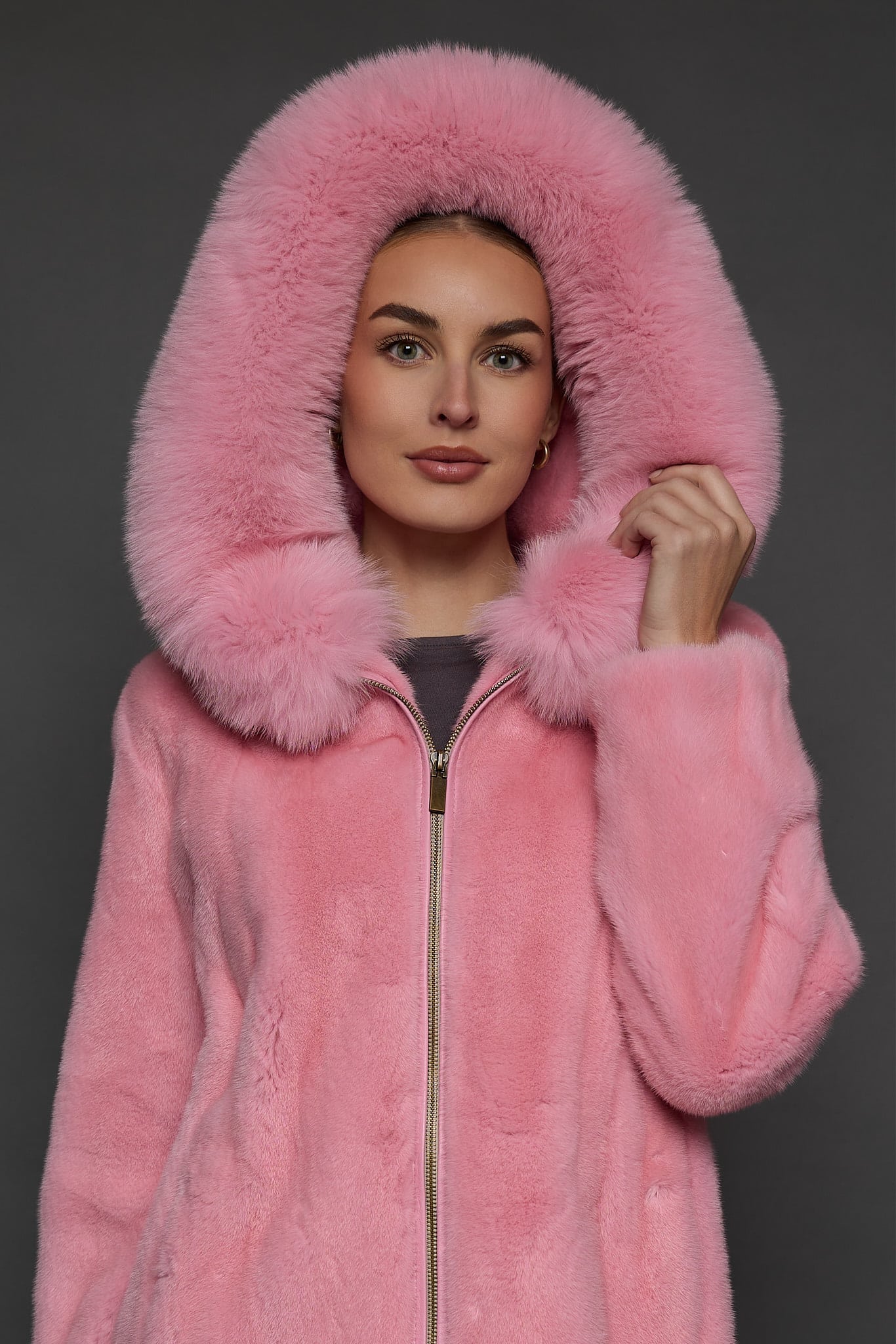 Close-up view of a woman in a blush mink jacket with the hood up standing in front of a dark grey background