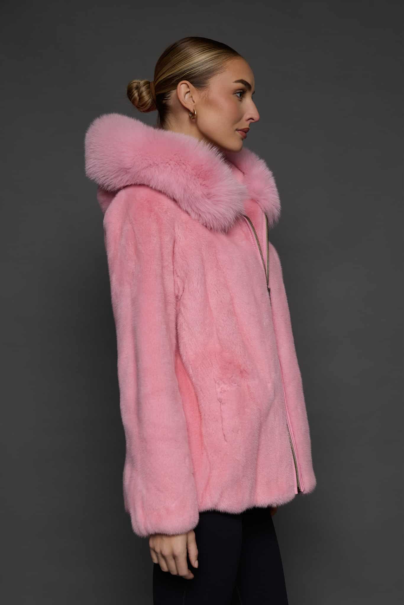 Profile view of a woman in a blush mink jacket with the hood down standing in front of a dark grey background