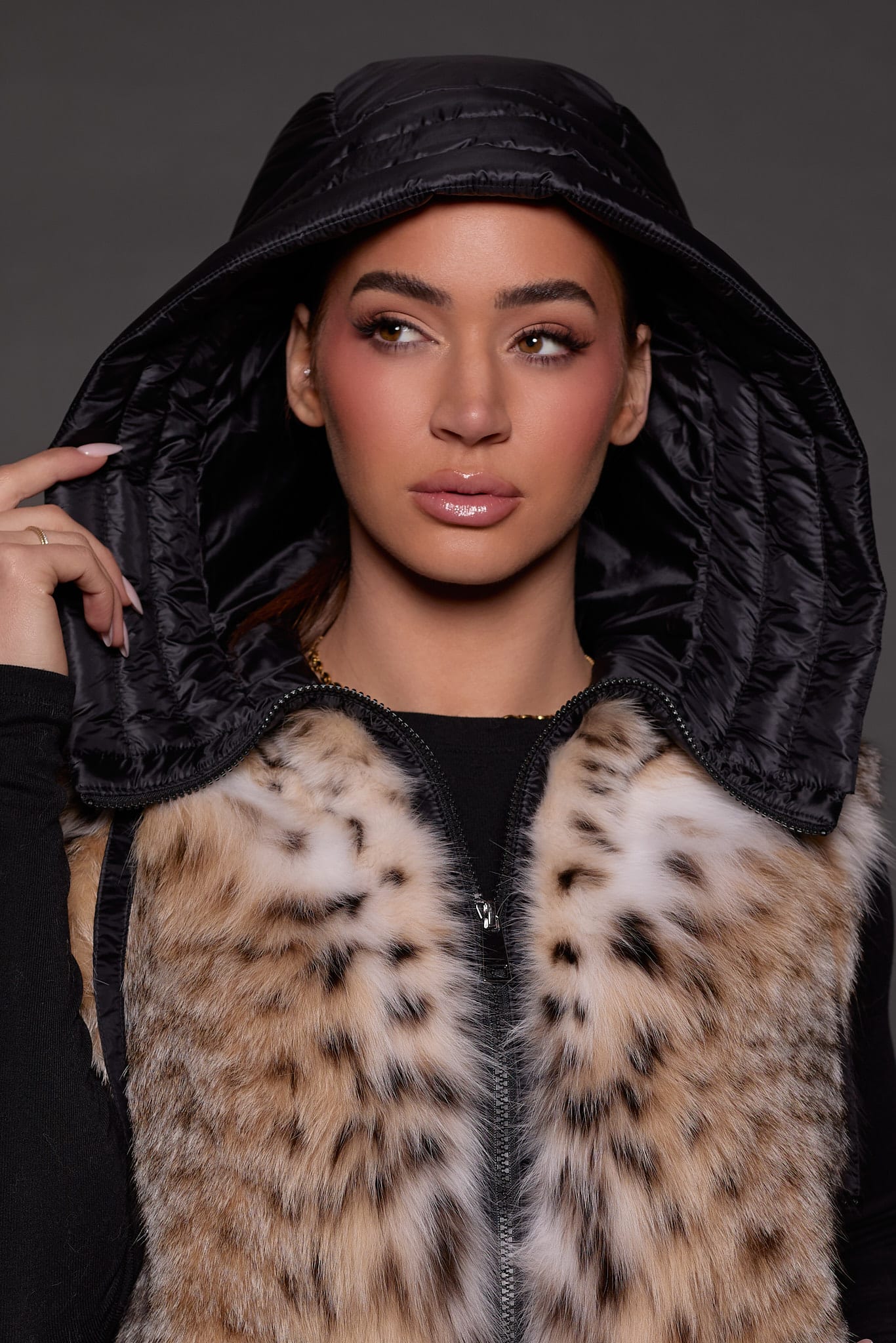 ANDI LYNX PUFFER VEST BLACK HOOD Up close image of a woman in a black and brown fur vest against a grey background with a black hood over her head
