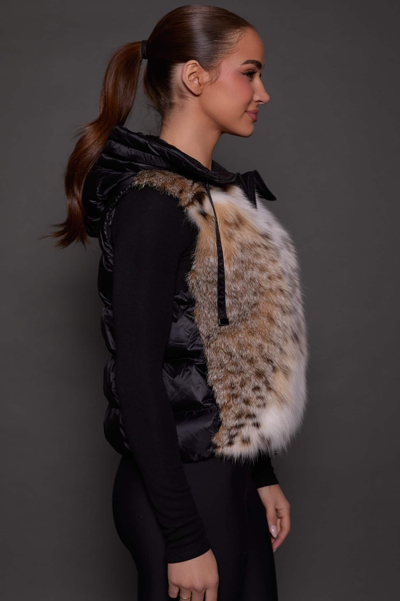 ANDI LYNX PUFFER VEST BLACK SIDE the side profile of a woman in a black and brown fur vest against a grey background