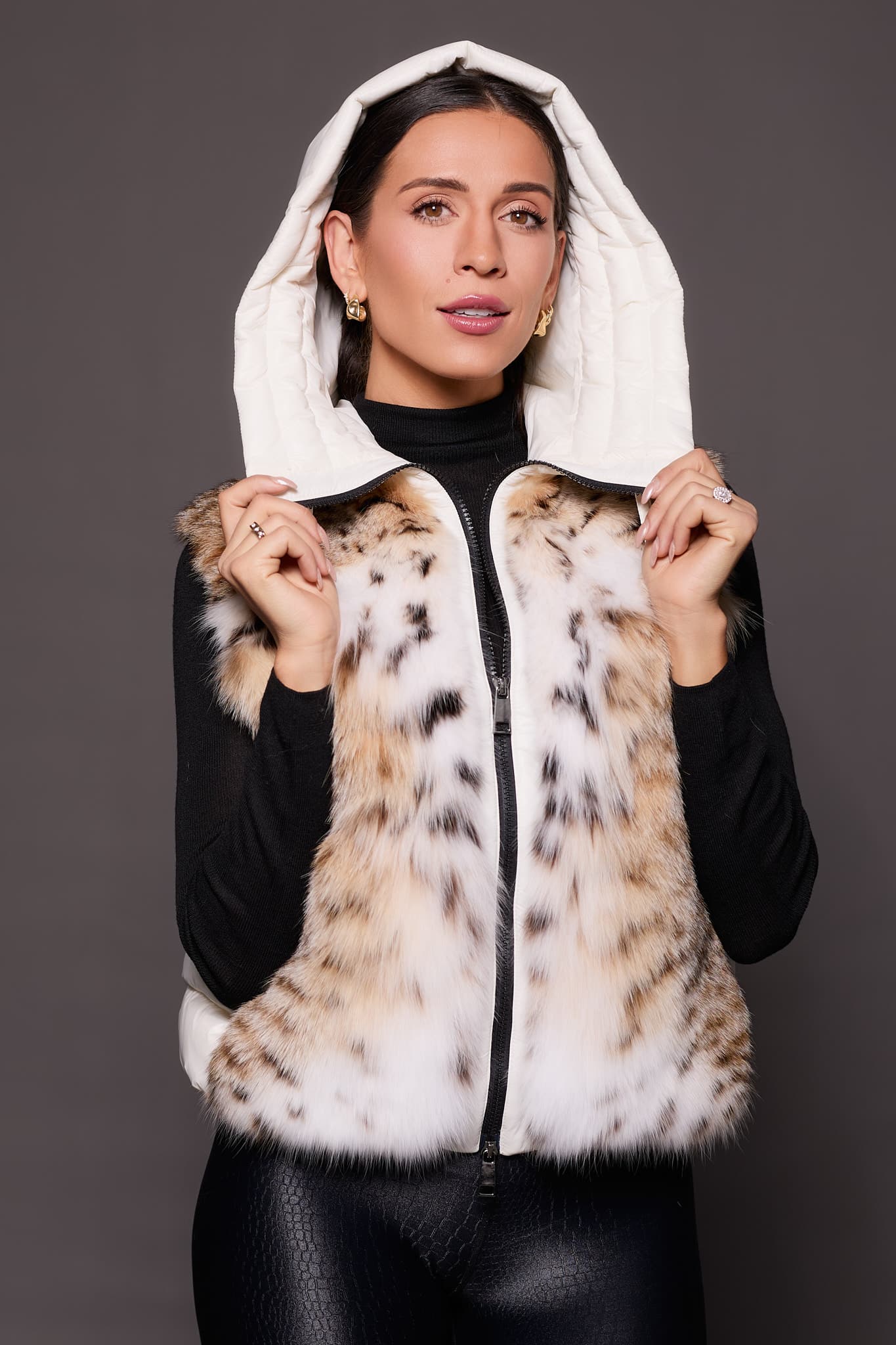 Andi Lynx Puffer Vest White Hood the front of a woman wearing a white puffer vest with lynx fur holding up the hood against a solid grey background