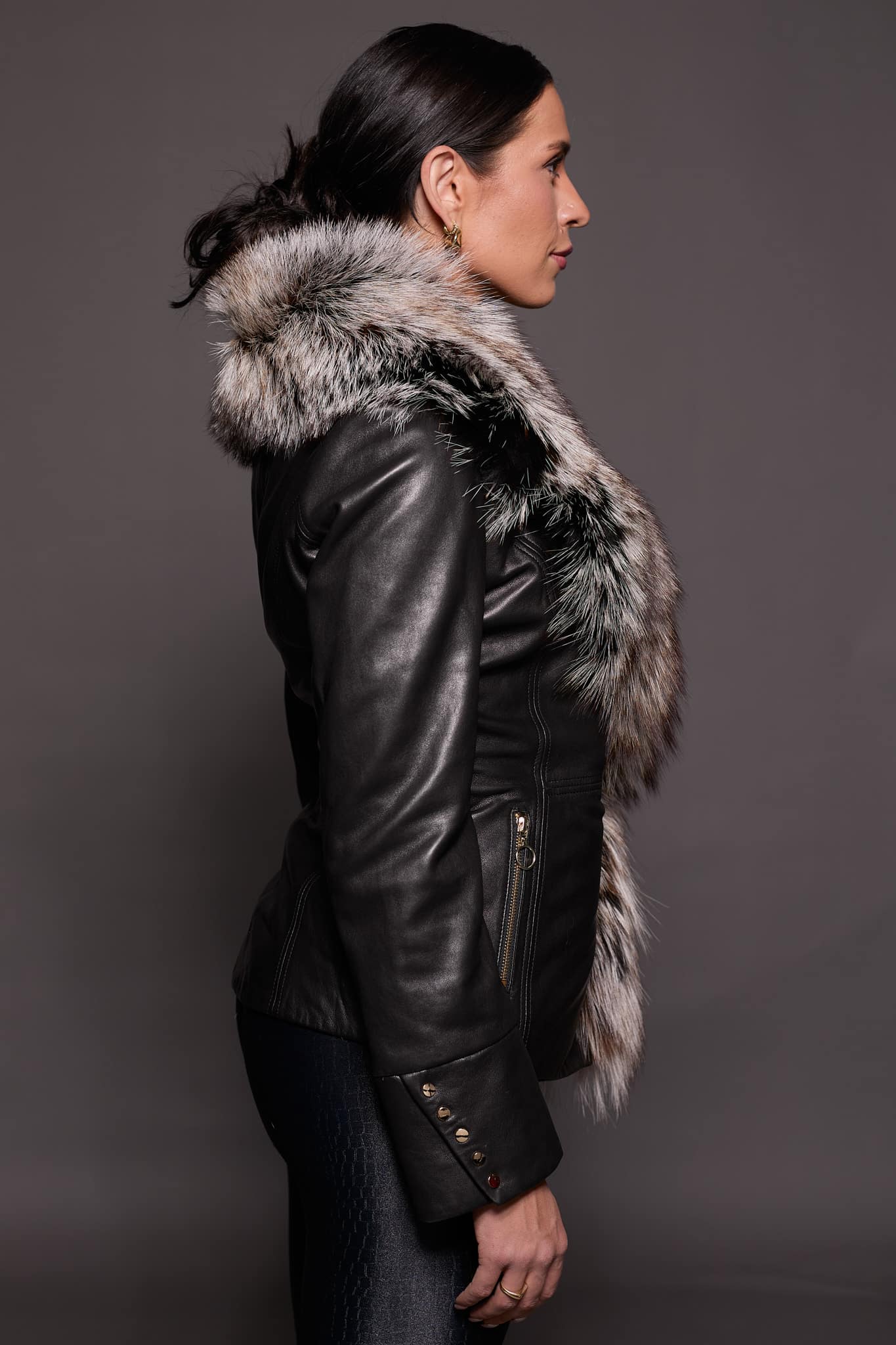The side profile of a woman in a black leather jacket with a brown fur trim against a dark grey background
