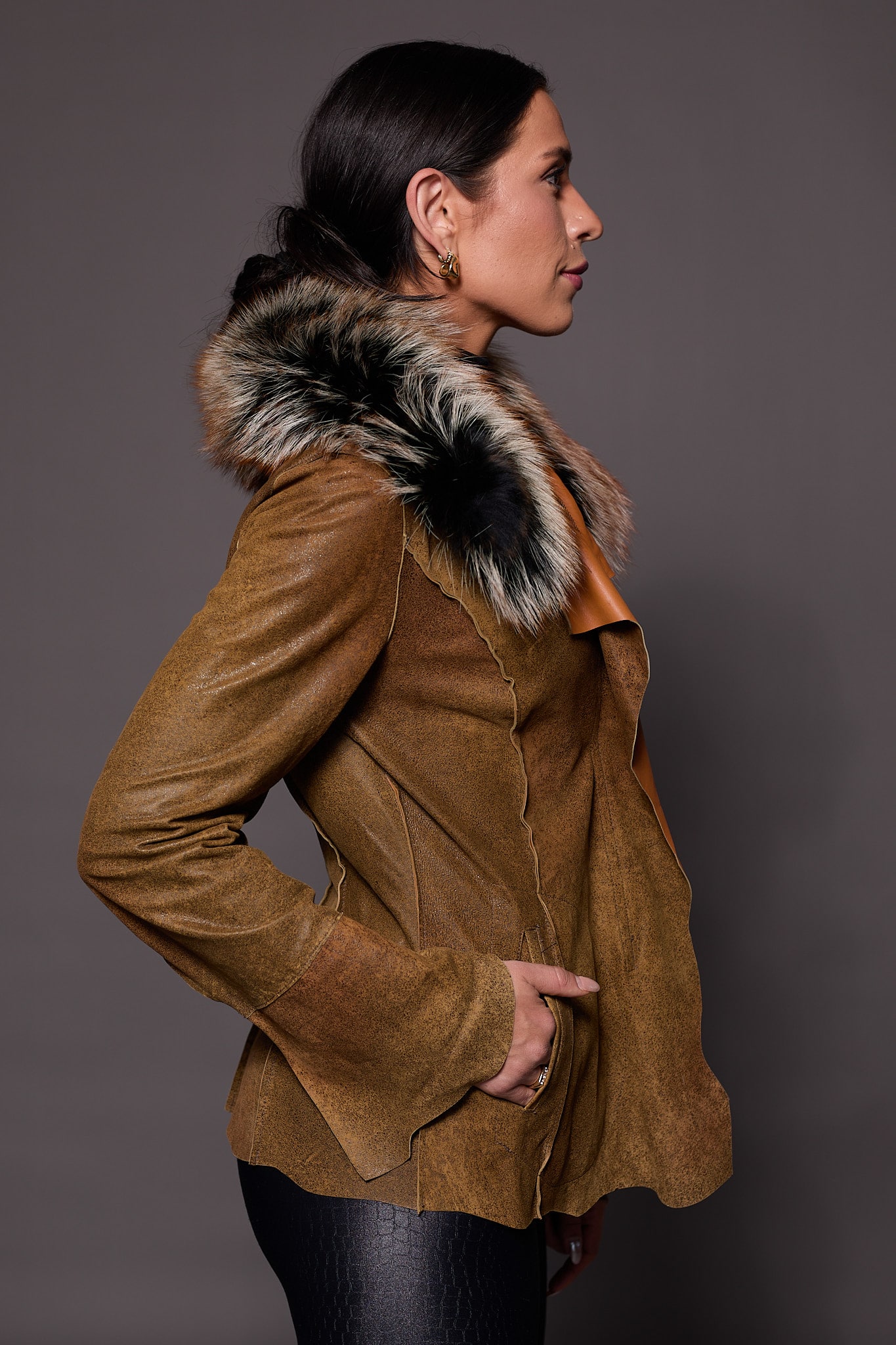 the side of a woman in a brown leather jacket with fur against a grey backdrop