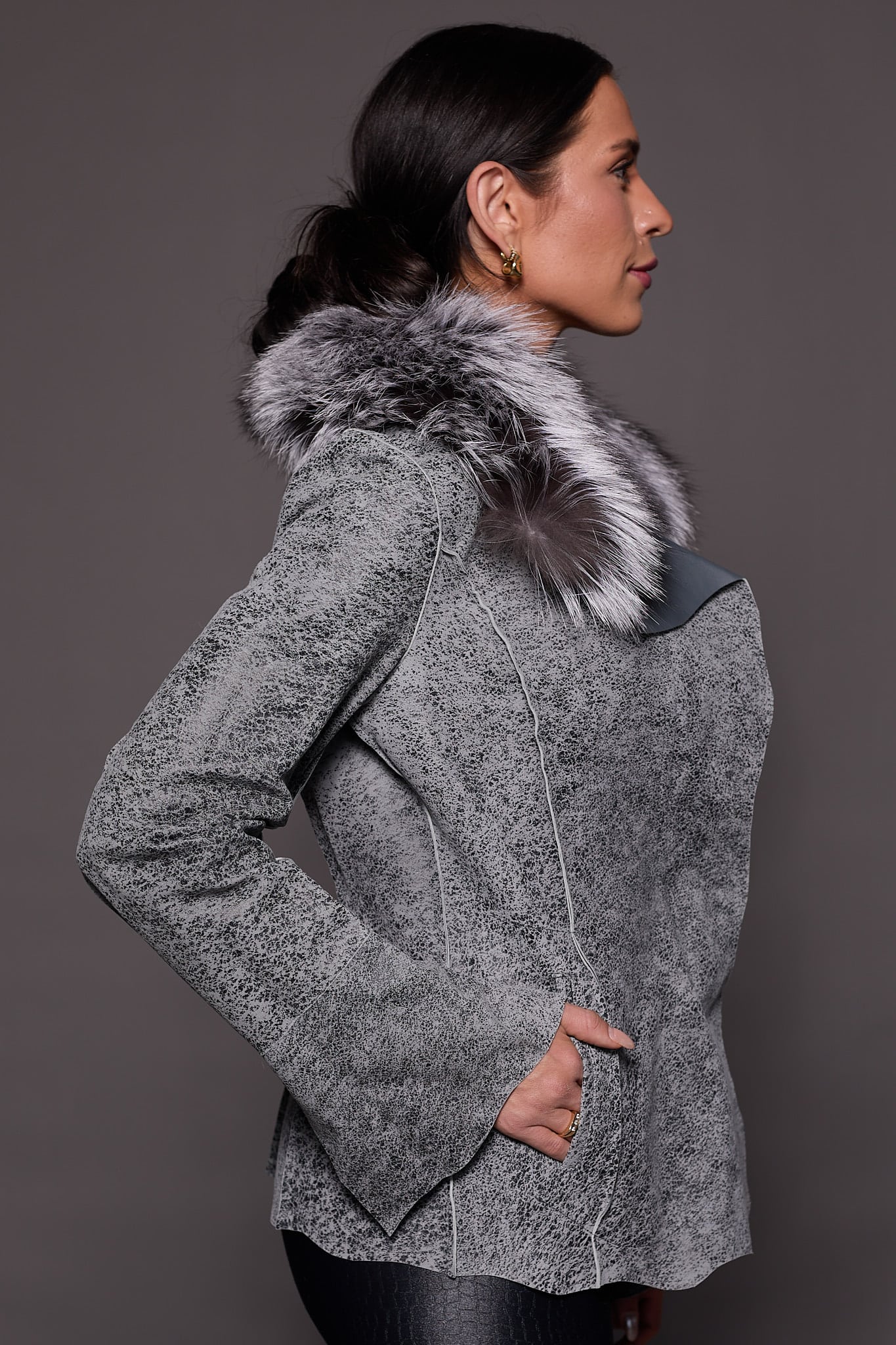 the side of a woman in a grey leather jacket with fur against a grey backdrop