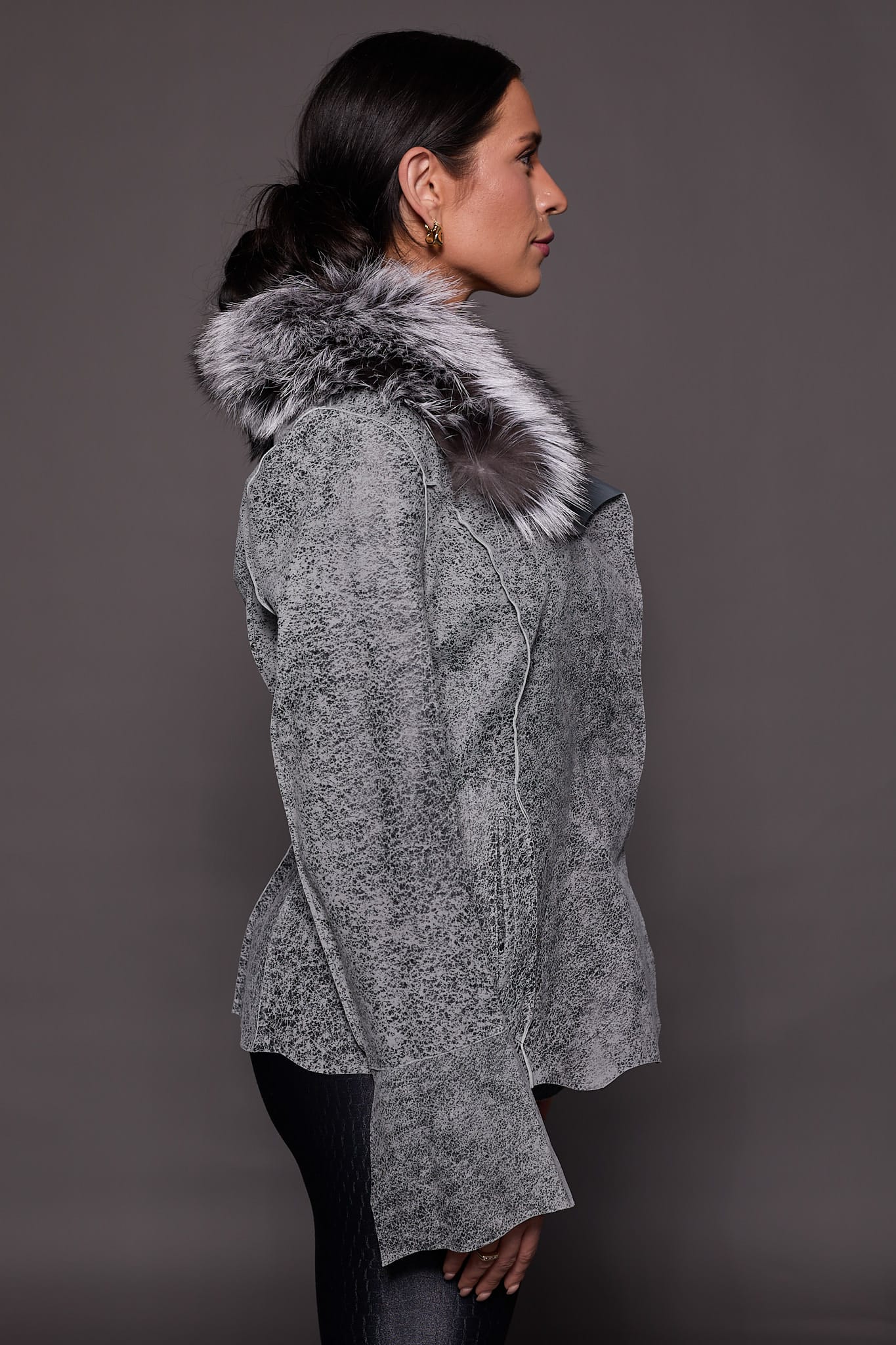 the side of a woman in a grey leather jacket with fur against a grey backdrop