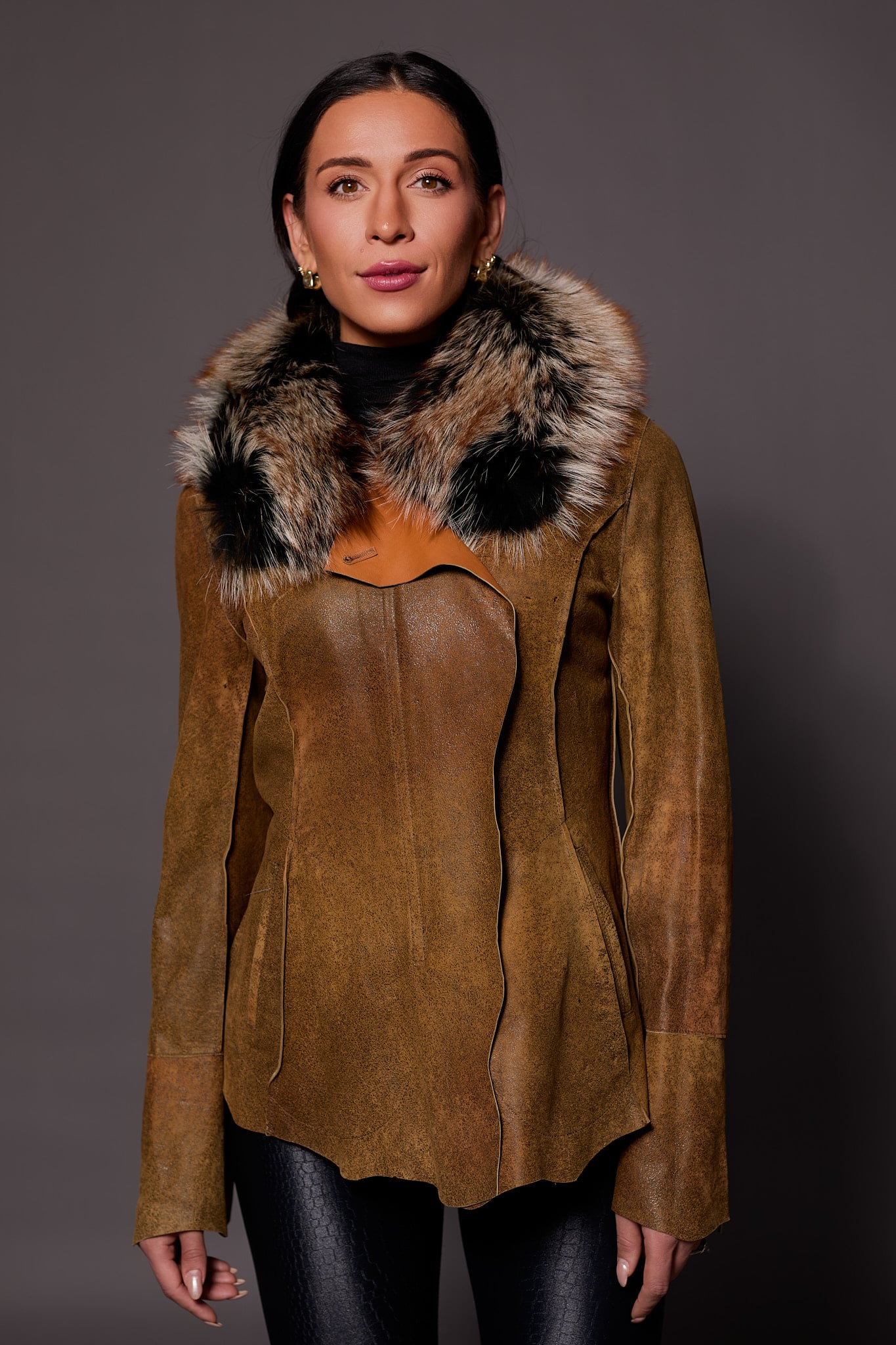 Frontal view of a woman in a brown leather jacket standing in front of a dark grey background.