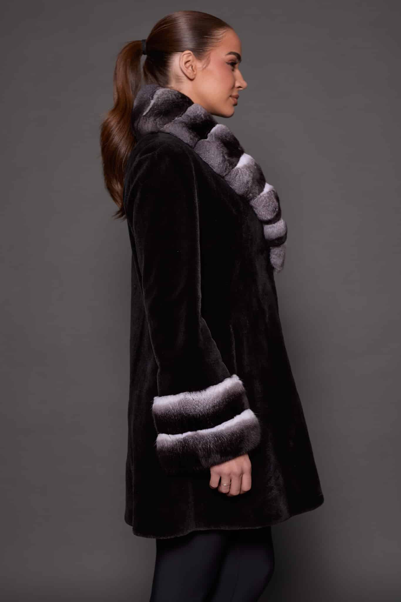 Profile view of a woman wearing a sheared mink fur coat standing in front of a grey background facing the right of frame.