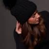 woman wearing a black knit beanie with a black fox puff on the top against a grey background