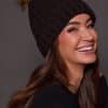 woman wearing a black knit beanie with a brown fox puff on the top against a grey background
