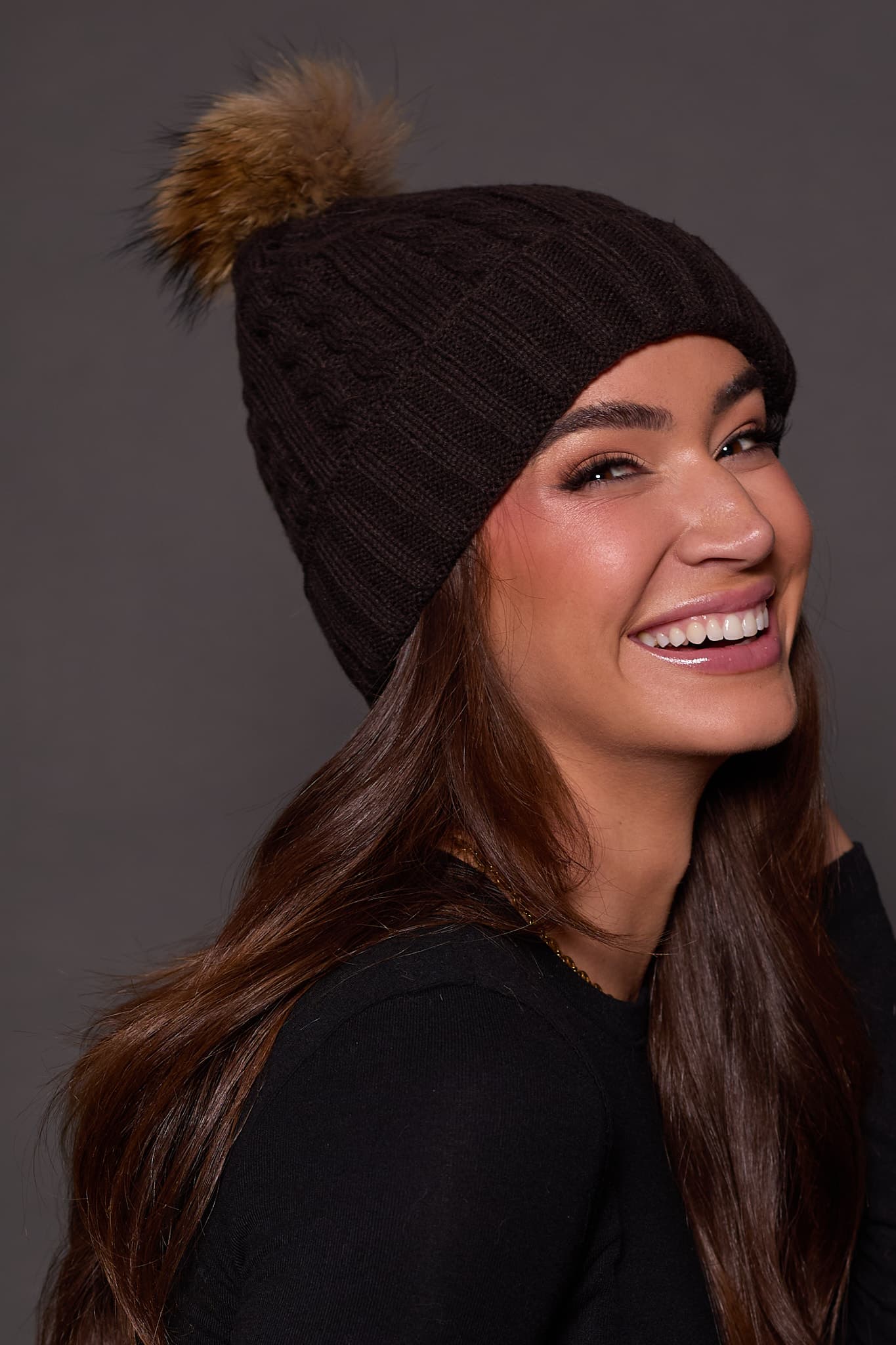 woman wearing a black knit beanie with a brown fox puff on the top against a grey background