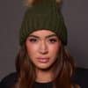 woman wearing a green knit beanie with a fox puff on the top against a grey background