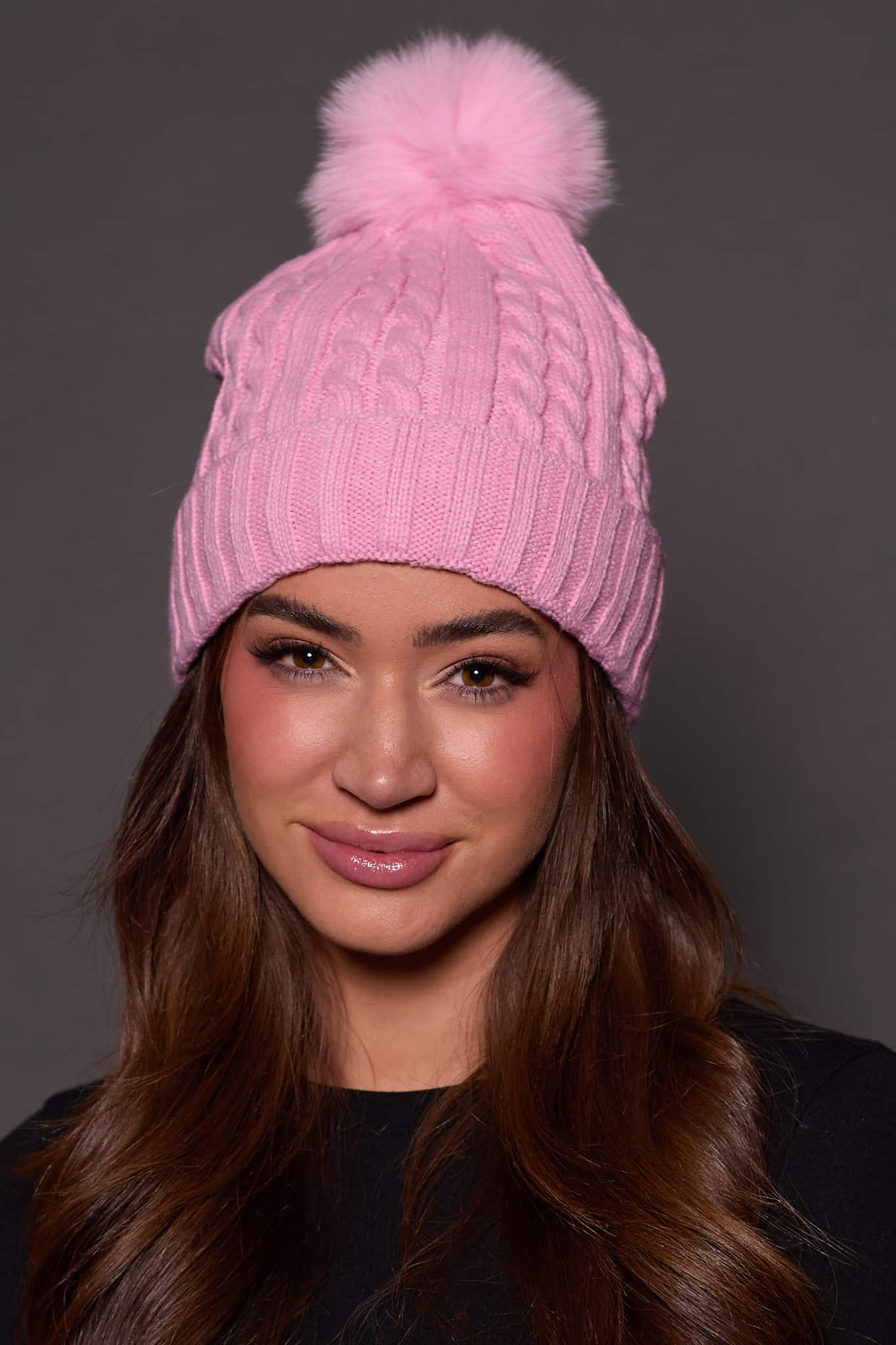 woman wearing a pink knit beanie with a fox puff on the top against a grey background