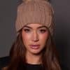 woman wearing a tan knit beanie with a fox puff on the top against a grey background