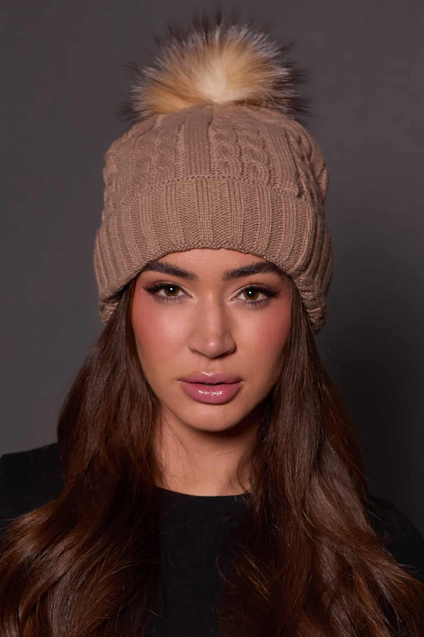 woman wearing a tan knit beanie with a fox puff on the top against a grey background