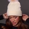 woman wearing a white knit beanie with a fox puff on the top against a grey background