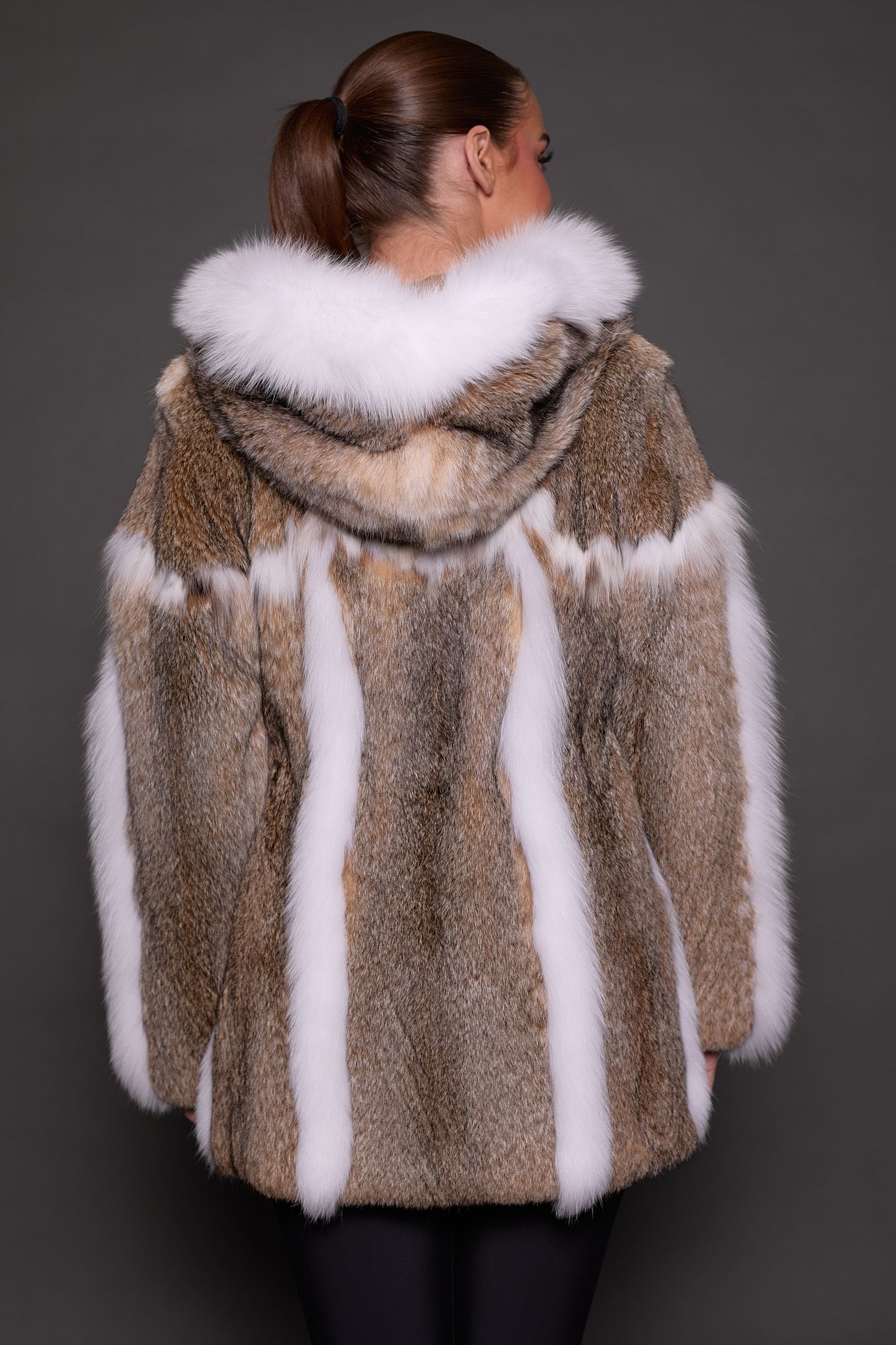 Rear view of a woman wearing a lynx fur coat with the hood down facing a grey background.