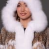 Close-up of a woman in a lynx fur coat with the hood up standing in front of a grey background.