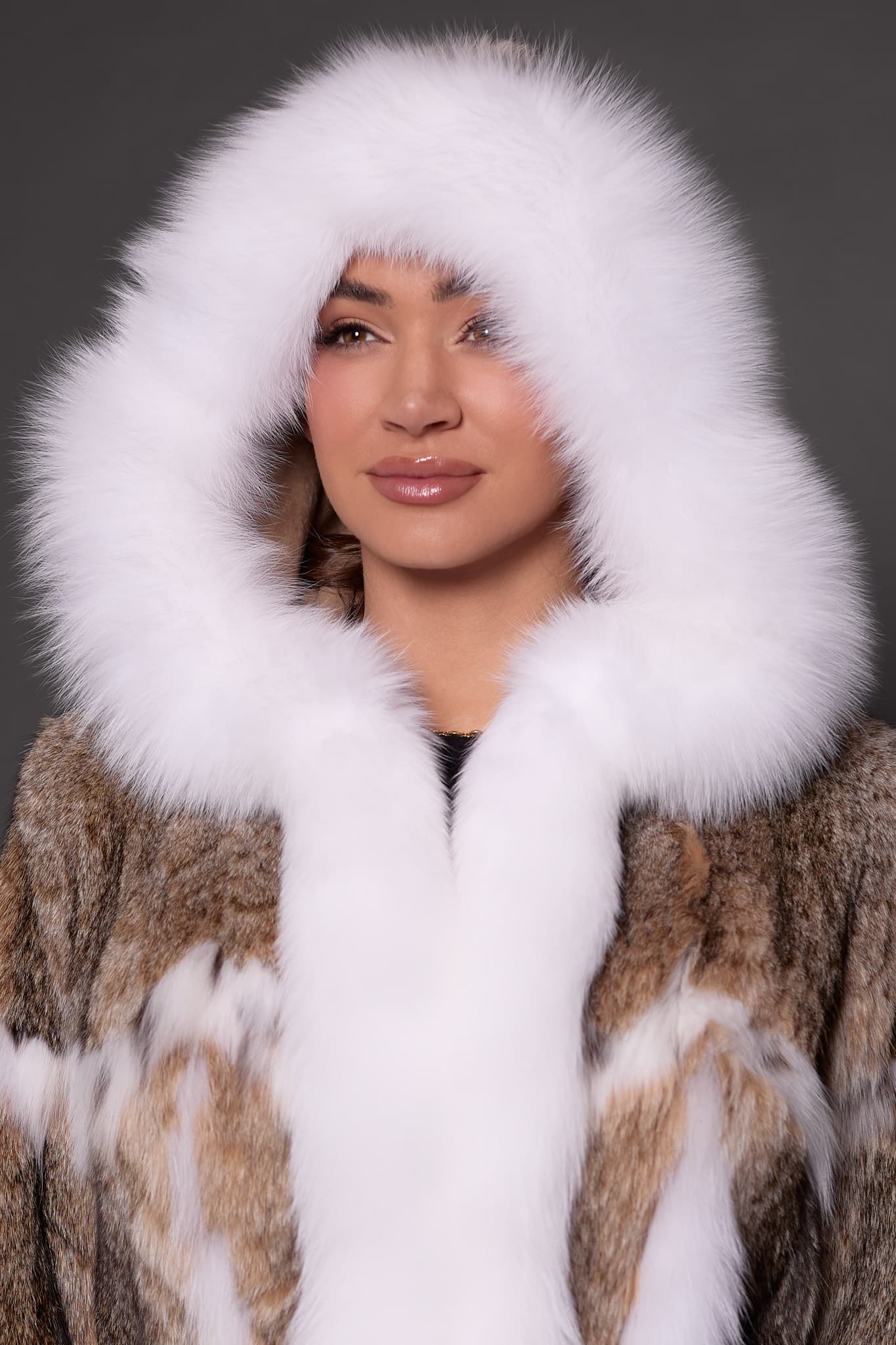Close-up of a woman in a lynx fur coat with the hood up standing in front of a grey background.