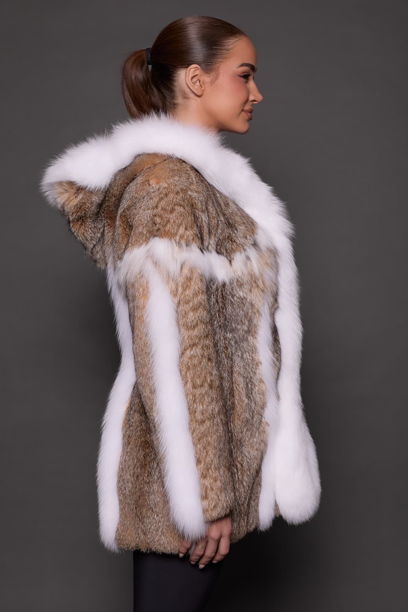 Profile view of a woman in a lynx fur coat with the hood down standing in front of a grey background. The woman is facing the right side of the frame.