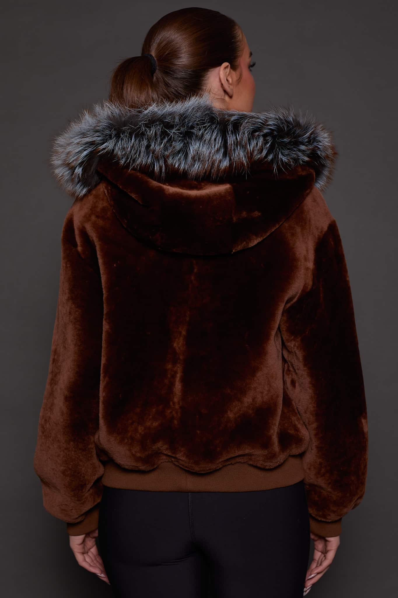 the back of a woman in a brown jacket with a silver fur trim on the hood against a grey background
