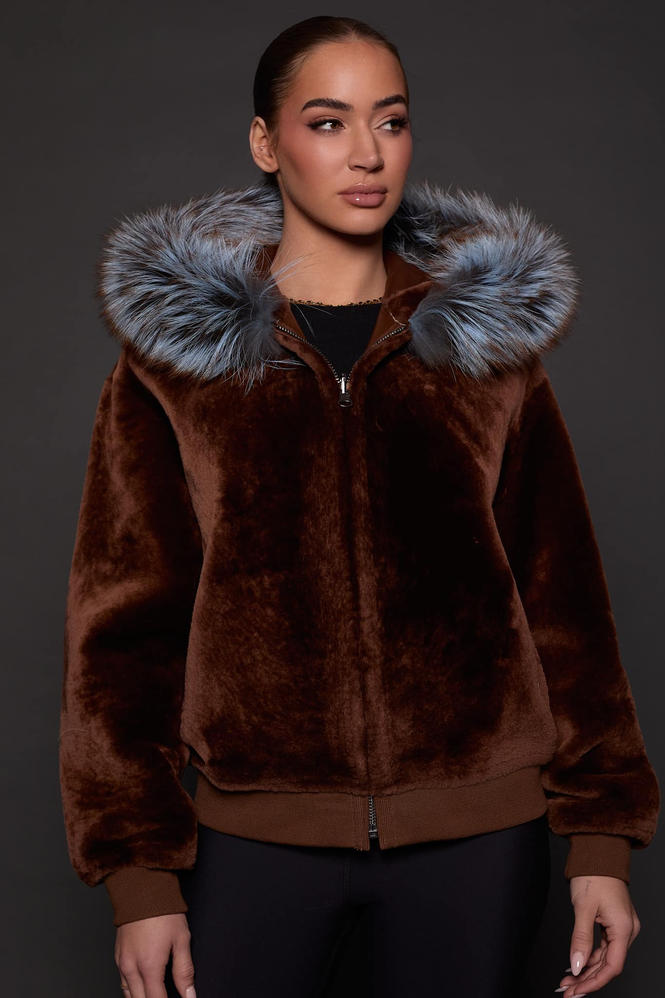 the front image of a woman in a brown jacket with a silver fur trim on the hood against a grey background