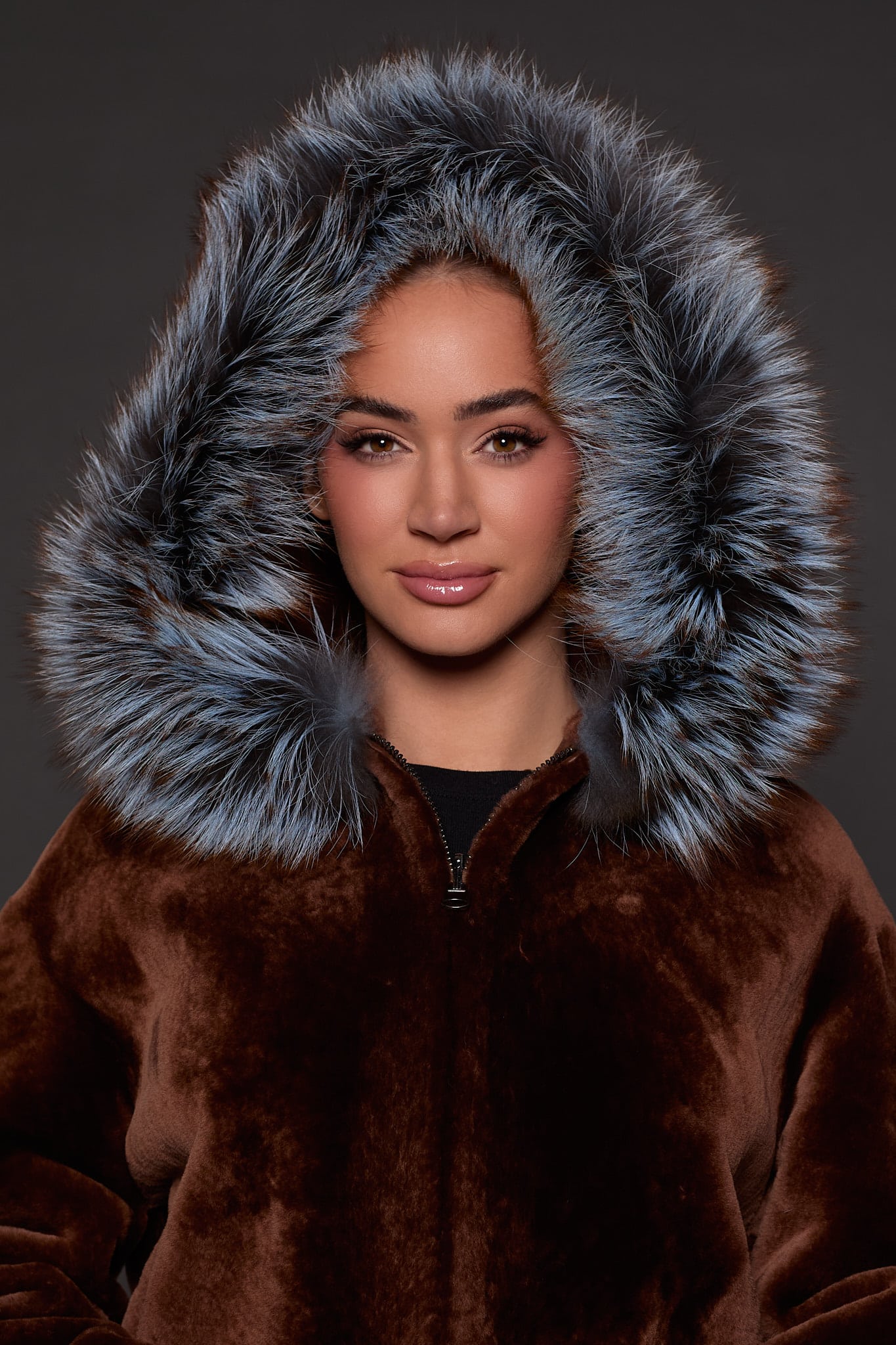 up close image of a woman in a brown jacket with a silver fur trim on the hood against a grey background