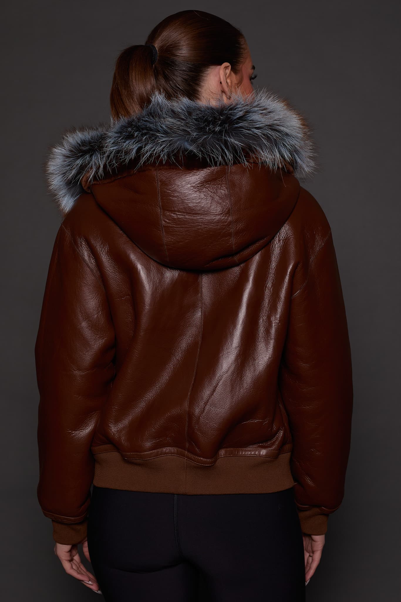 the back image of a woman in a brown jacket with a silver fur trim on the hood against a grey background
