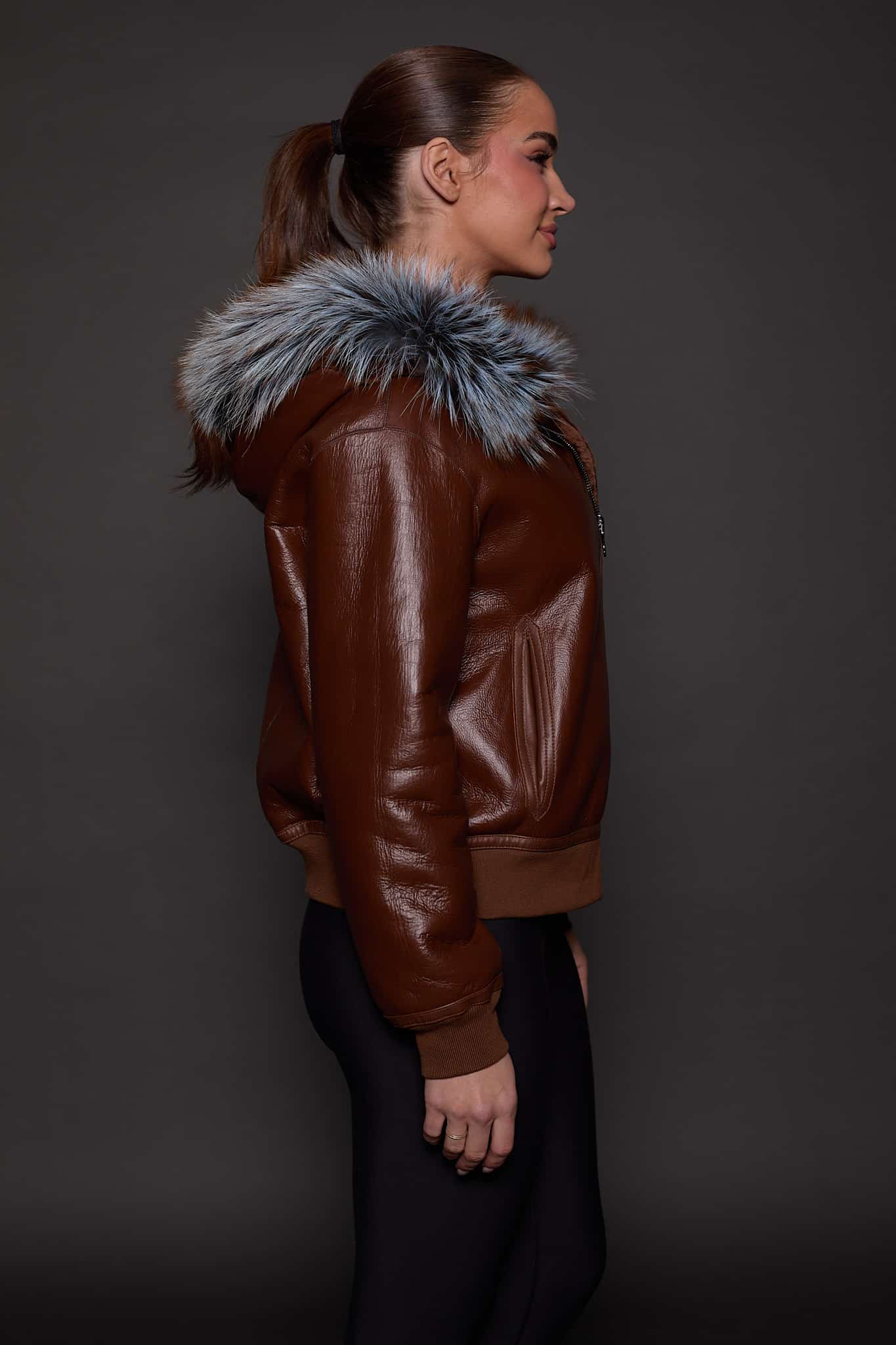 the side profile of a woman in a brown jacket with a silver fur trim on the hood against a grey background