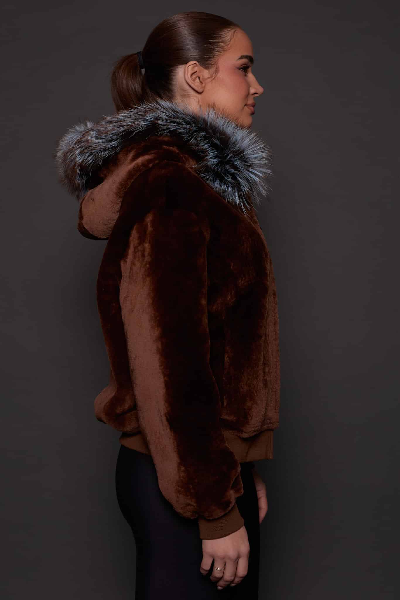 the side profile of a woman in a brown jacket with a silver fur trim on the hood against a grey background