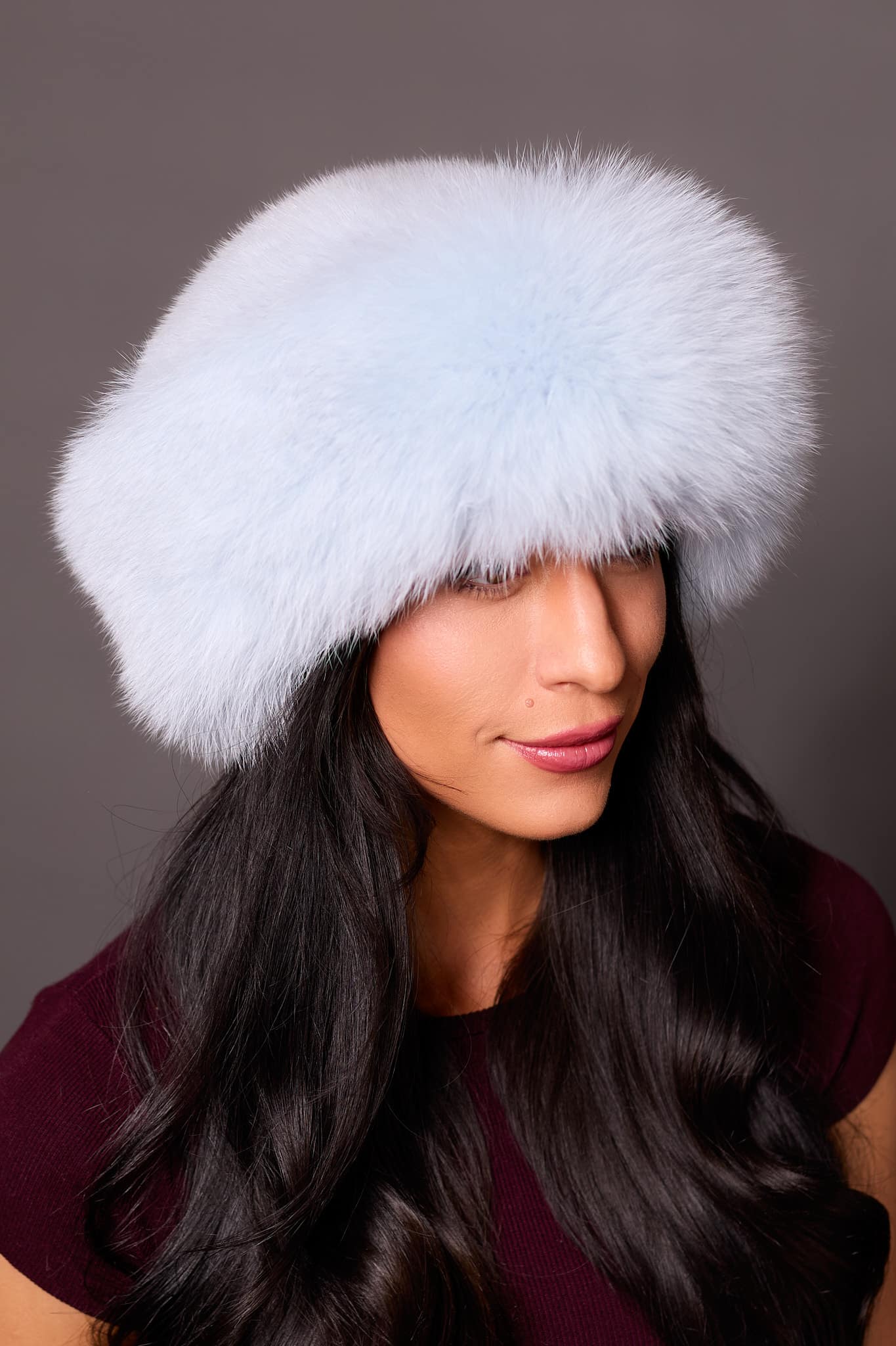 High angle frontal view of a woman in a baby blue mink fur and baby blue fox fur hat standing in front of a grey background.