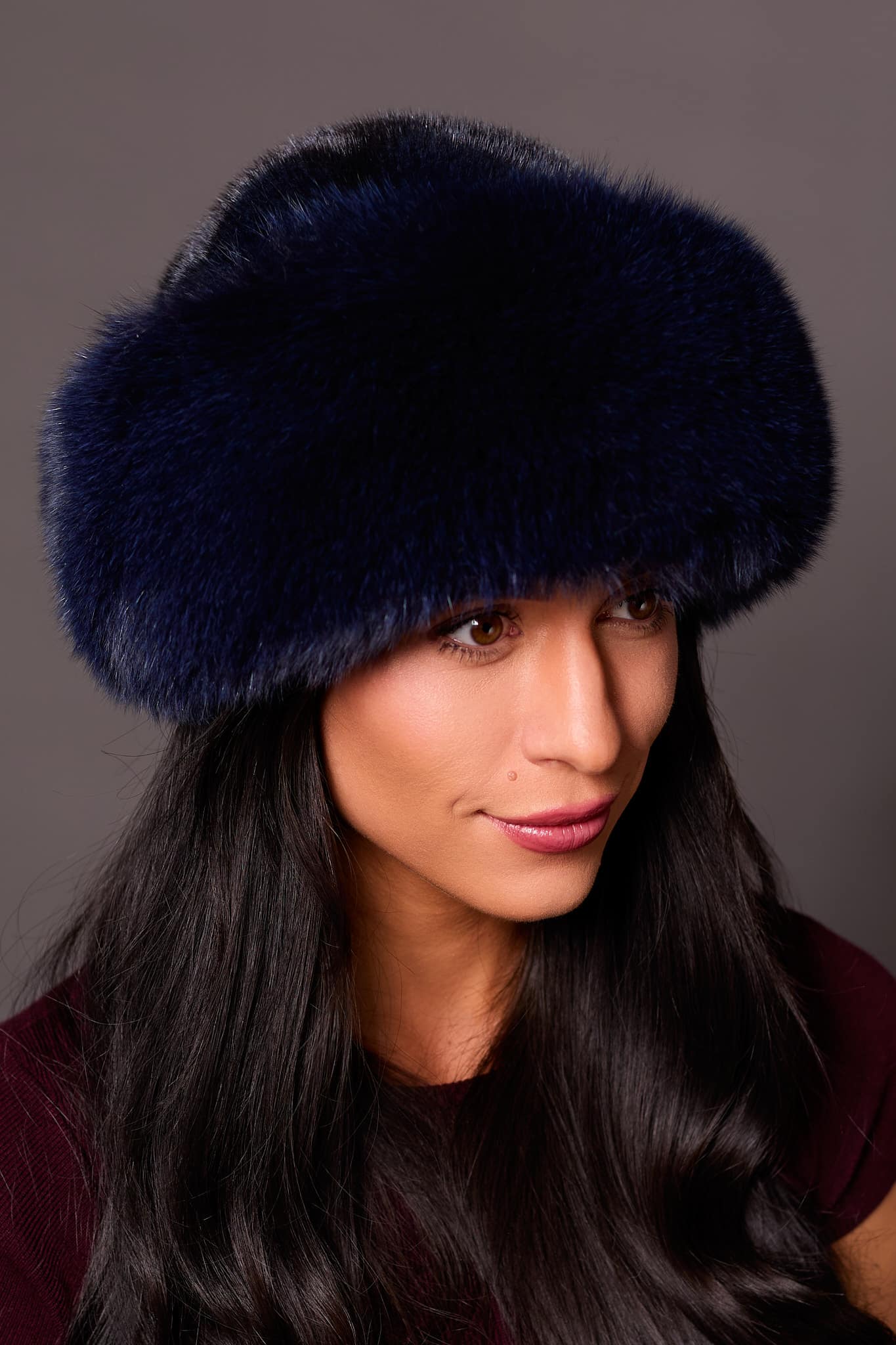 High angle frontal view of a woman in a blue mink fur and blue fox fur hat standing in front of a grey background.