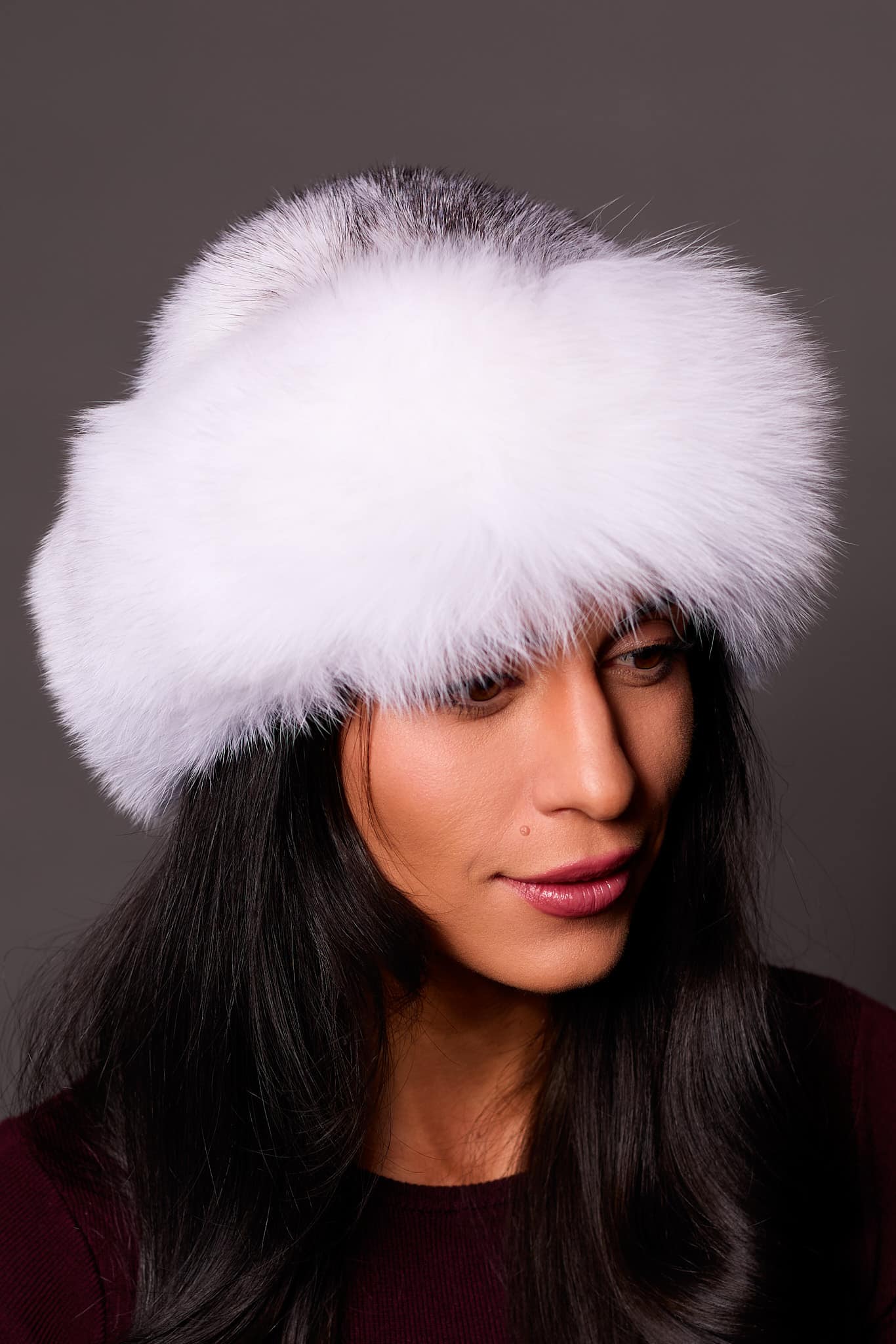 High angle frontal view of a woman in a cross mink fur and arctic fox fur hat standing in front of a grey background.