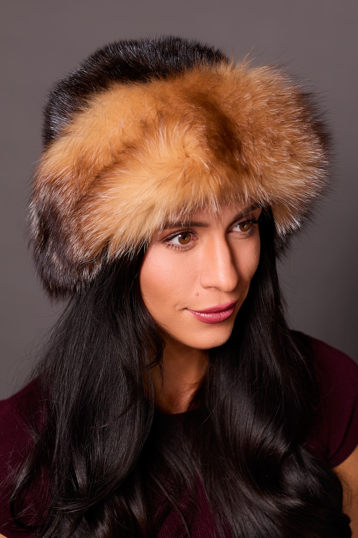 High angle frontal view of a woman in a mahogany mink fur and crystal fox fur hat standing in front of a grey background.