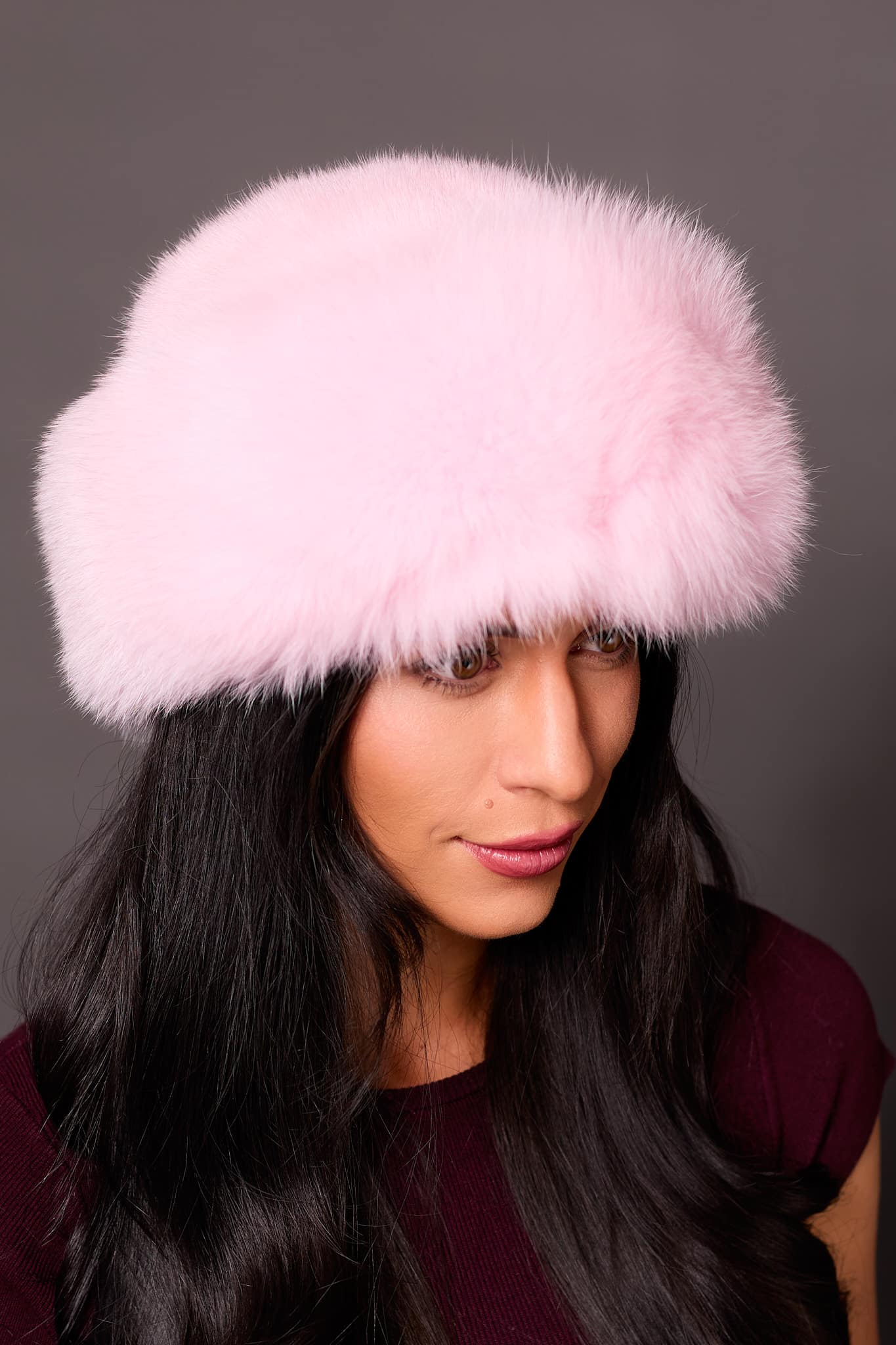 High angle frontal view of a woman in a pink mink fur and pink fox fur hat standing in front of a grey background.