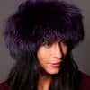 High angle frontal view of a woman in a purple mink fur and purple fox fur hat standing in front of a grey background.