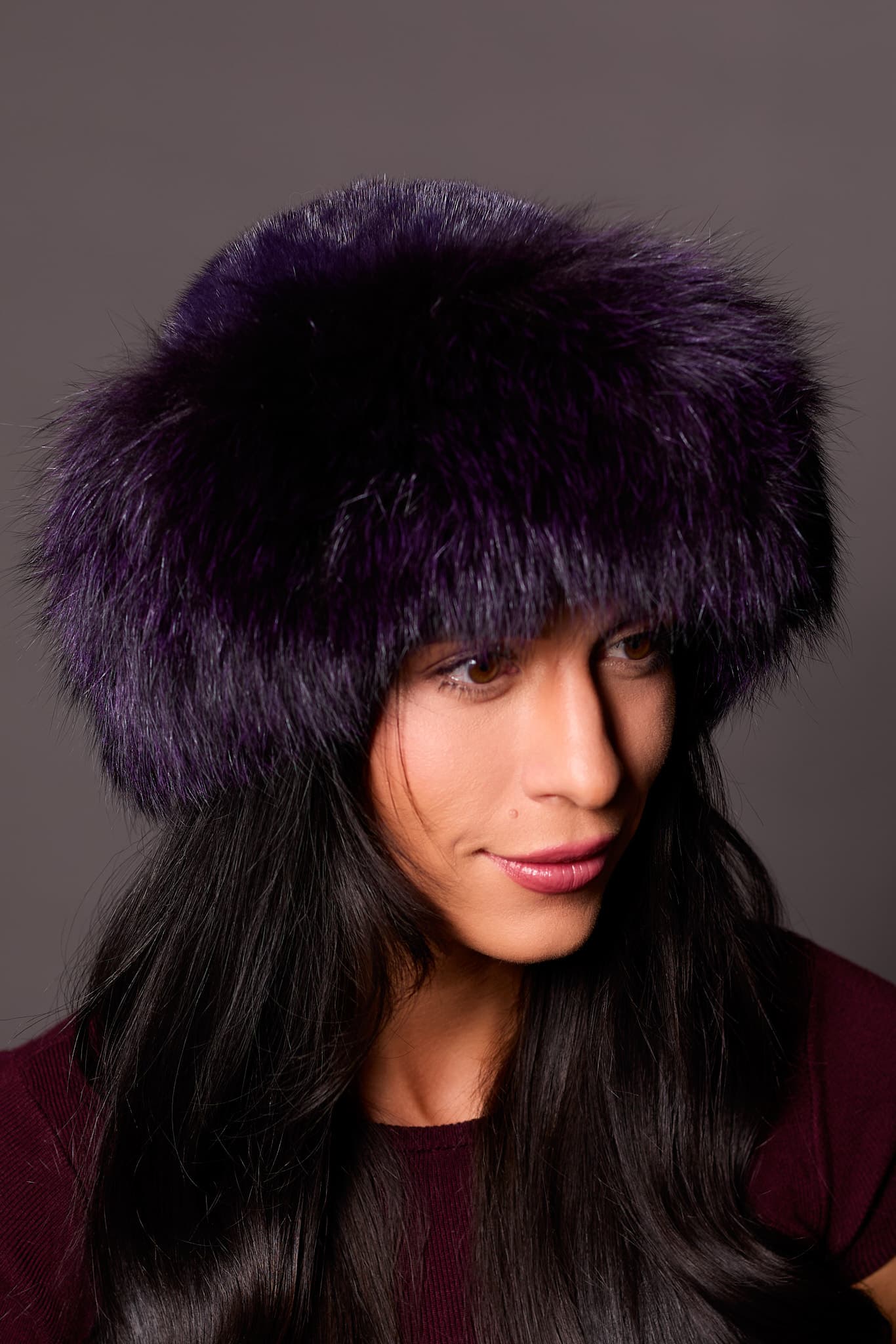 High angle frontal view of a woman in a purple mink fur and purple fox fur hat standing in front of a grey background.