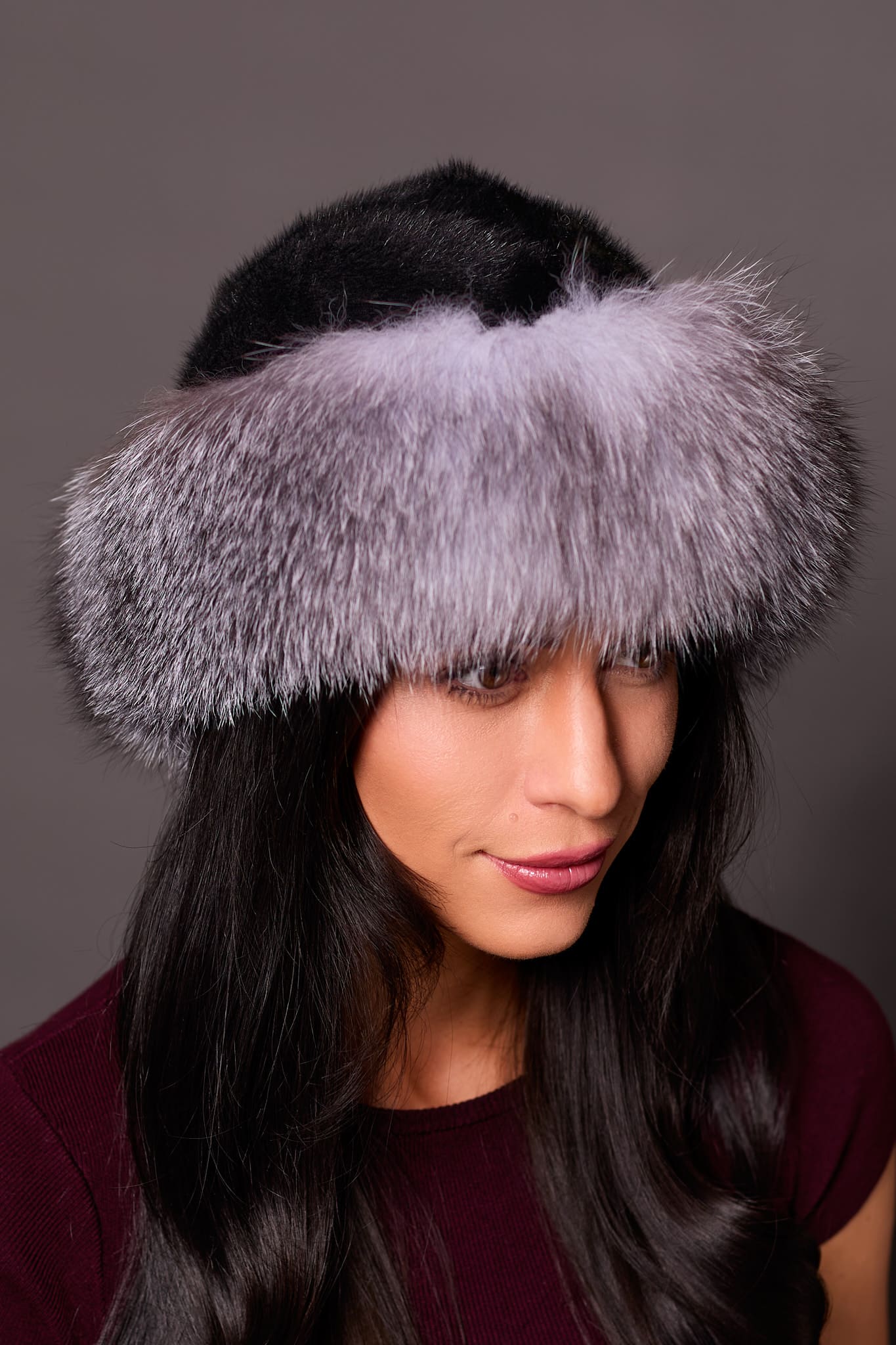 High angle frontal view of a woman in a ranch mink fur and indigo fox fur hat standing in front of a grey background.