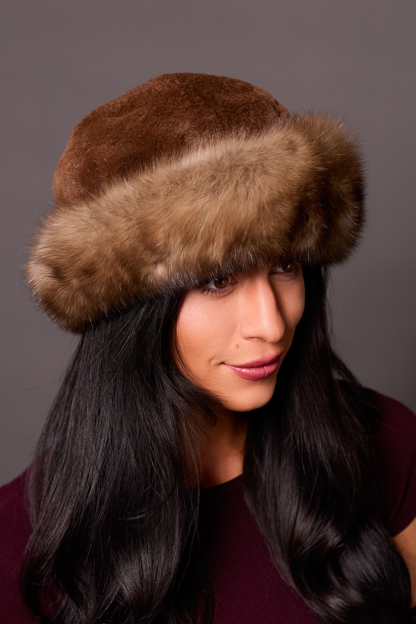 High angle frontal view of a woman wearing a sheared mink fur and Russian sable fur hat standing in front of a grey background.