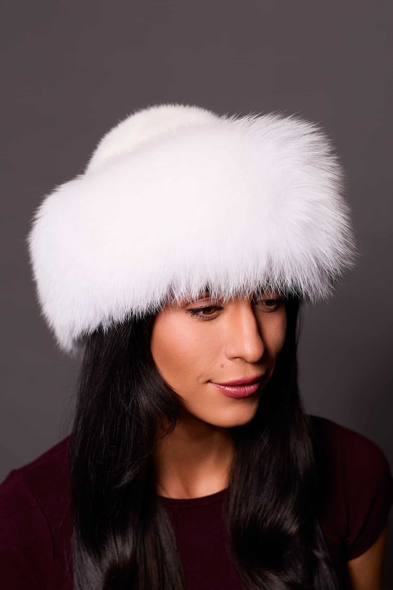 High angle frontal view of a woman in a white mink fur and arctic fox fur hat standing in front of a grey background.