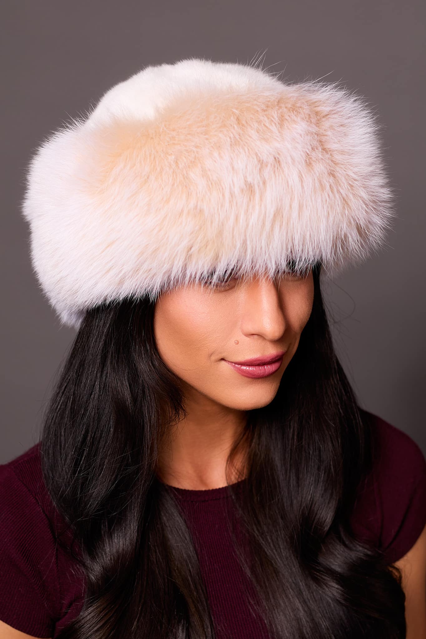High angle frontal view of a woman in a white mink fur and blush fox fur hat standing in front of a grey background.